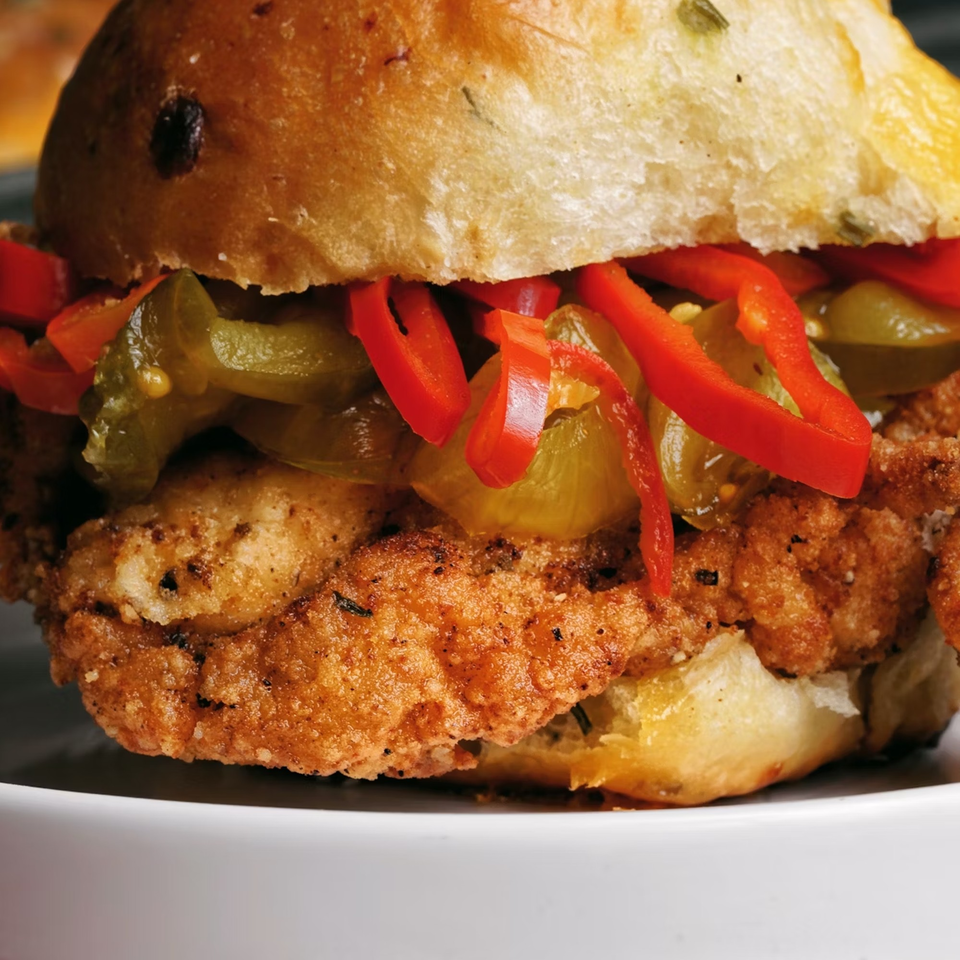 Close-up of a fried chicken sandwich with red peppers and pickled jalapeños on a bun.