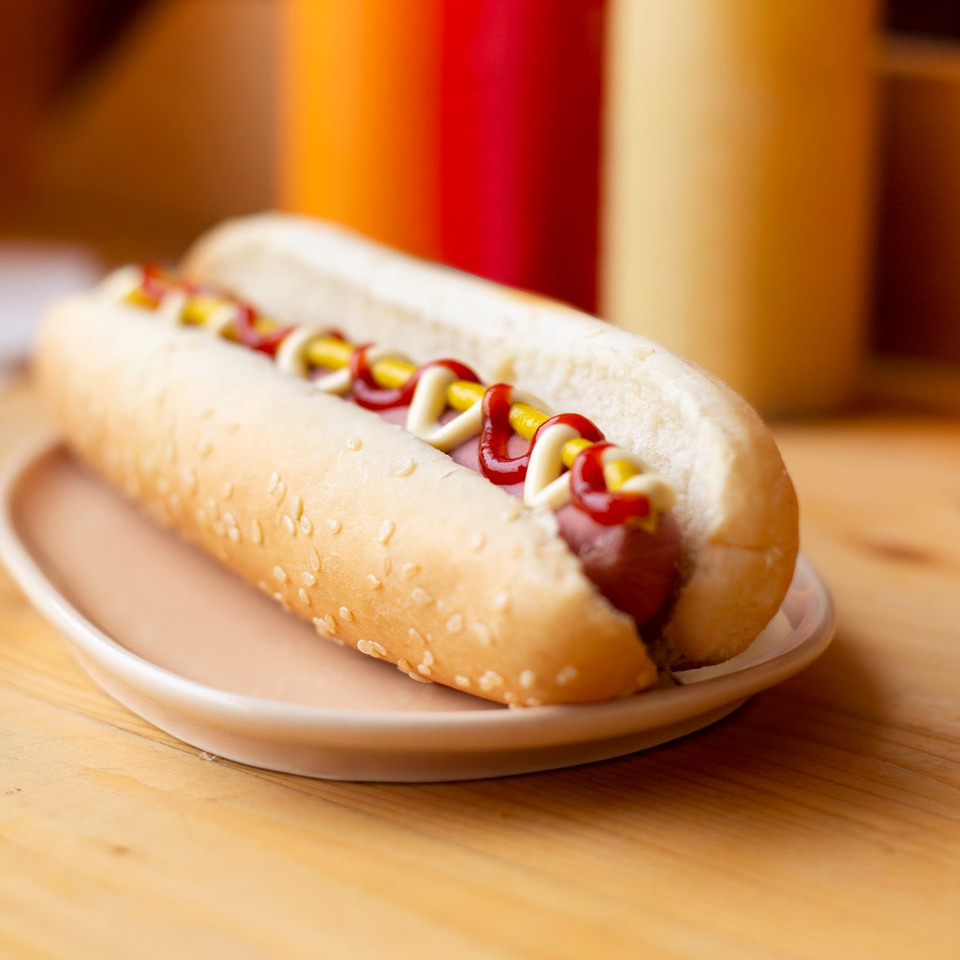 A hot dog with ketchup, mustard, and mayonnaise on a sesame seed bun, on a plate.
