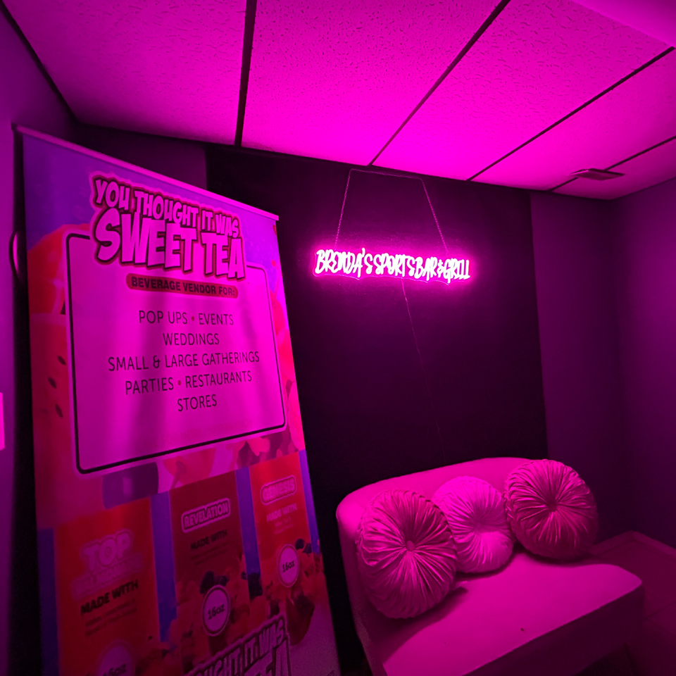 Brenda's Sports Bar & Grill neon sign glows pink above a white couch with round pillows.