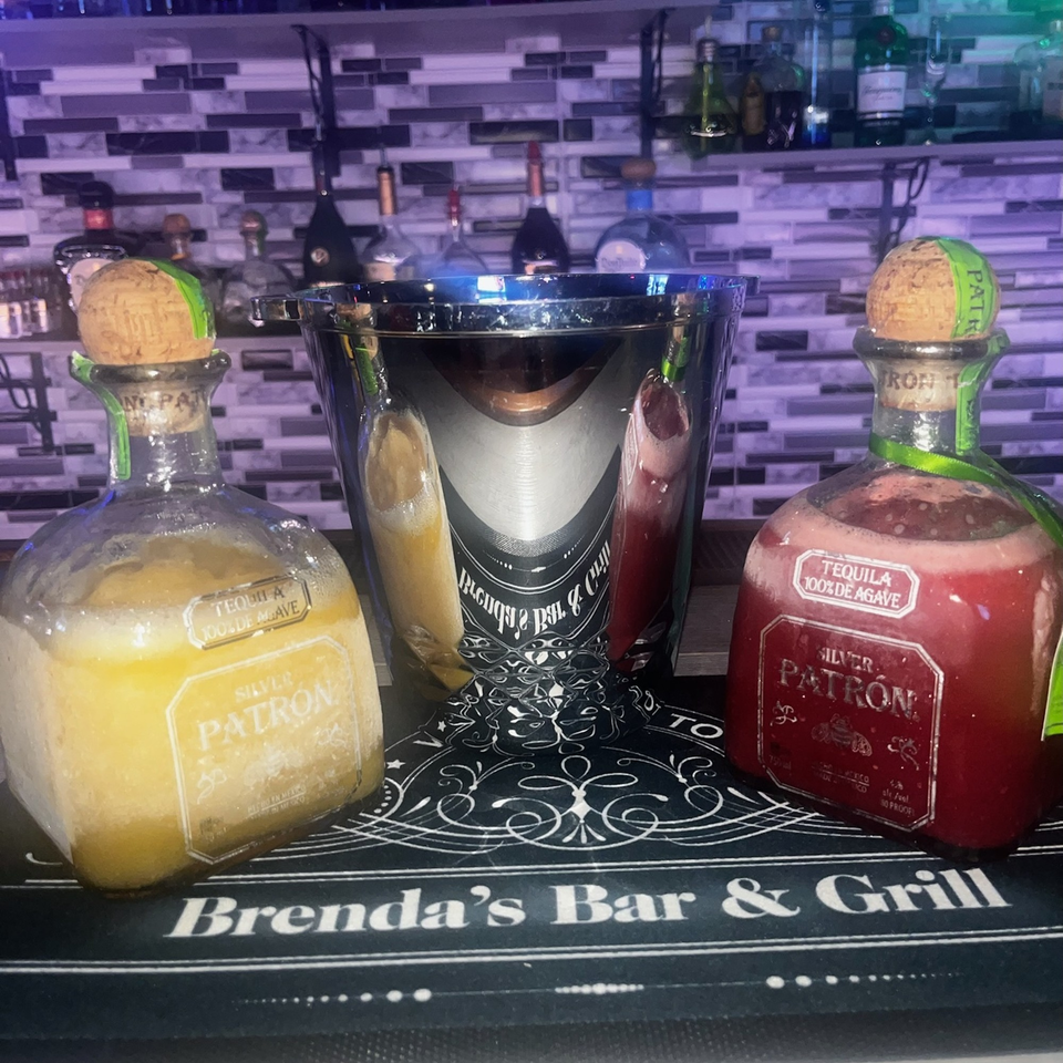 Two Patron tequila bottles, one yellow and one red, flank a silver ice bucket on a bar mat.