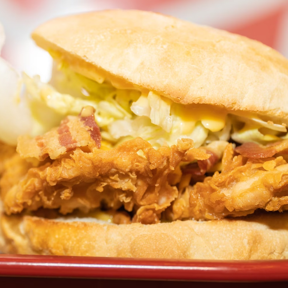 Close-up of a crispy fried chicken sandwich with bacon, lettuce, and sauce on a red plate.