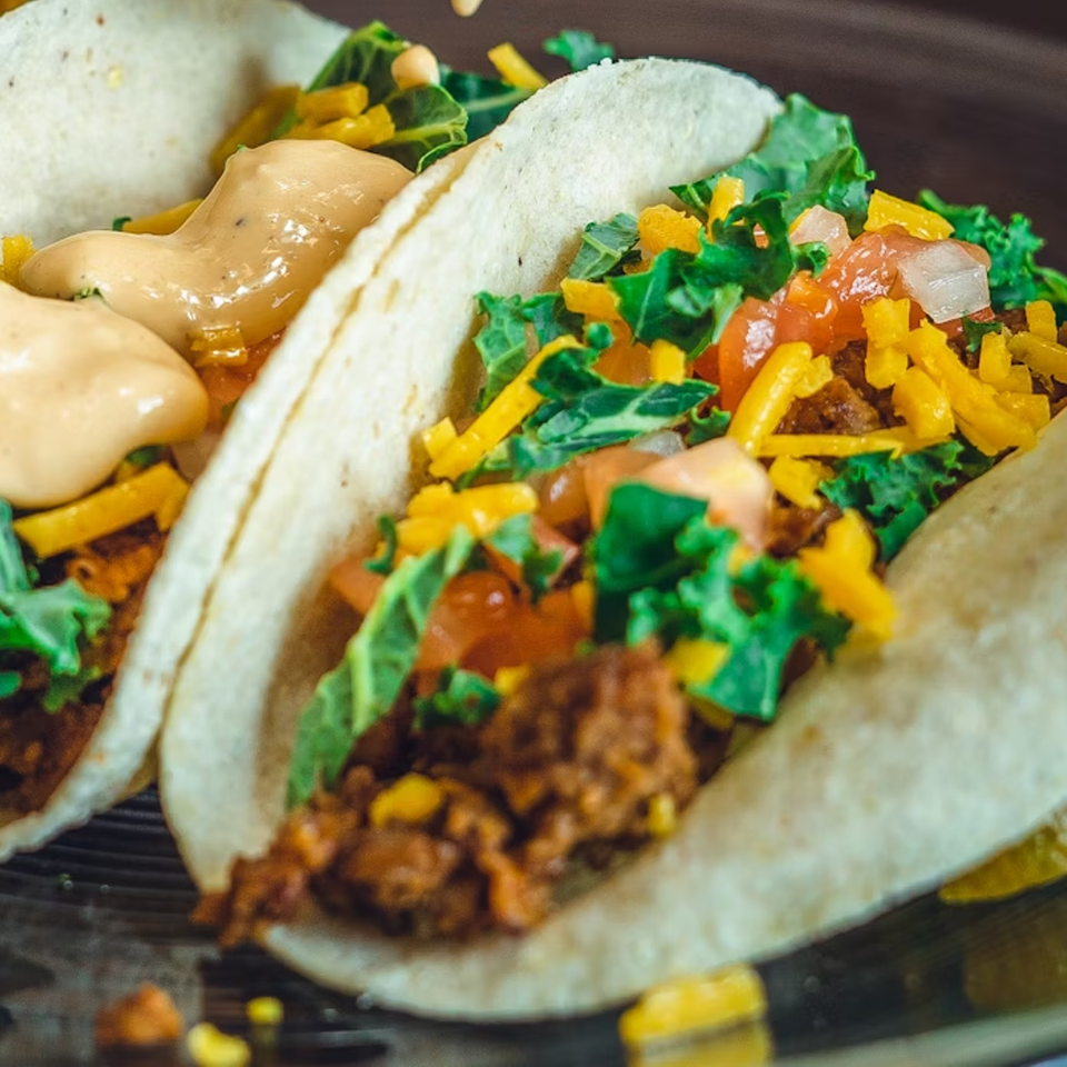 Two soft tacos filled with seasoned meat, shredded cheese, lettuce, diced tomatoes, and creamy sauce.