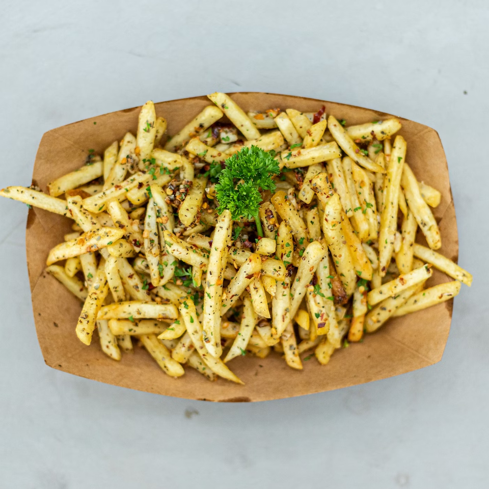 A brown paper tray filled with golden french fries, seasoned with herbs and spices, garnished with parsley.
