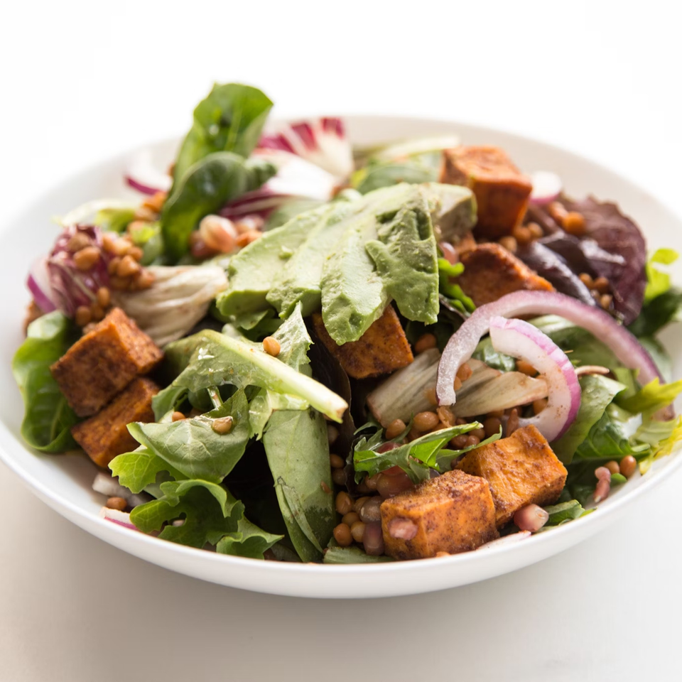 A vibrant salad with greens, avocado, sweet potato, lentils, and red onion in a white bowl.